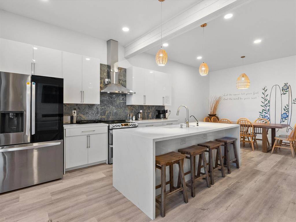 This modern kitchen features sleek white cabinetry, stainless steel appliances, and a marble backsplash. A large island with wooden stools provides seating, and pendant lighting adds warmth. The adjacent dining area includes a wooden table and chairs, complemented by decorative wall art.