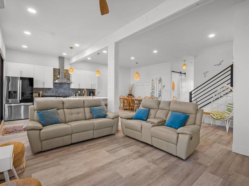 Bright, open-concept living area with modern kitchen, featuring stainless steel appliances and sleek cabinetry. The space includes comfortable seating and a dining area, complemented by stylish lighting and wood flooring. A staircase leads to additional levels, adding to the spacious feel.