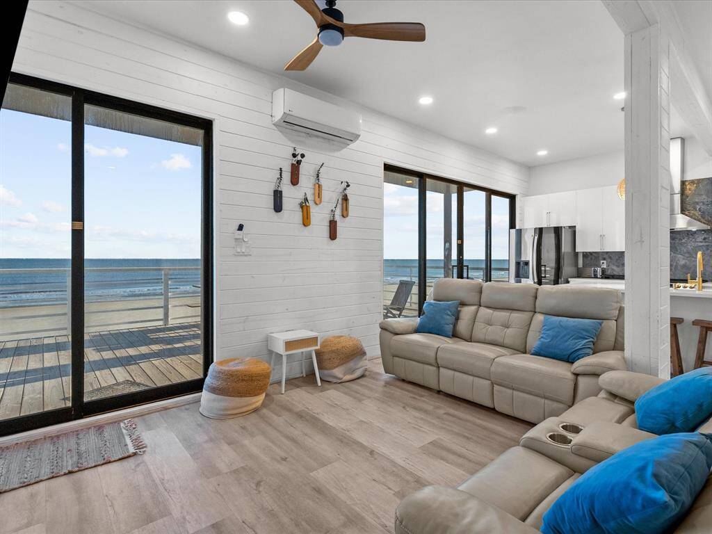 Bright and modern beachfront living room with ocean views, featuring large sliding glass doors, comfortable seating, wood accents, and an open-plan layout leading to a stylish kitchen.