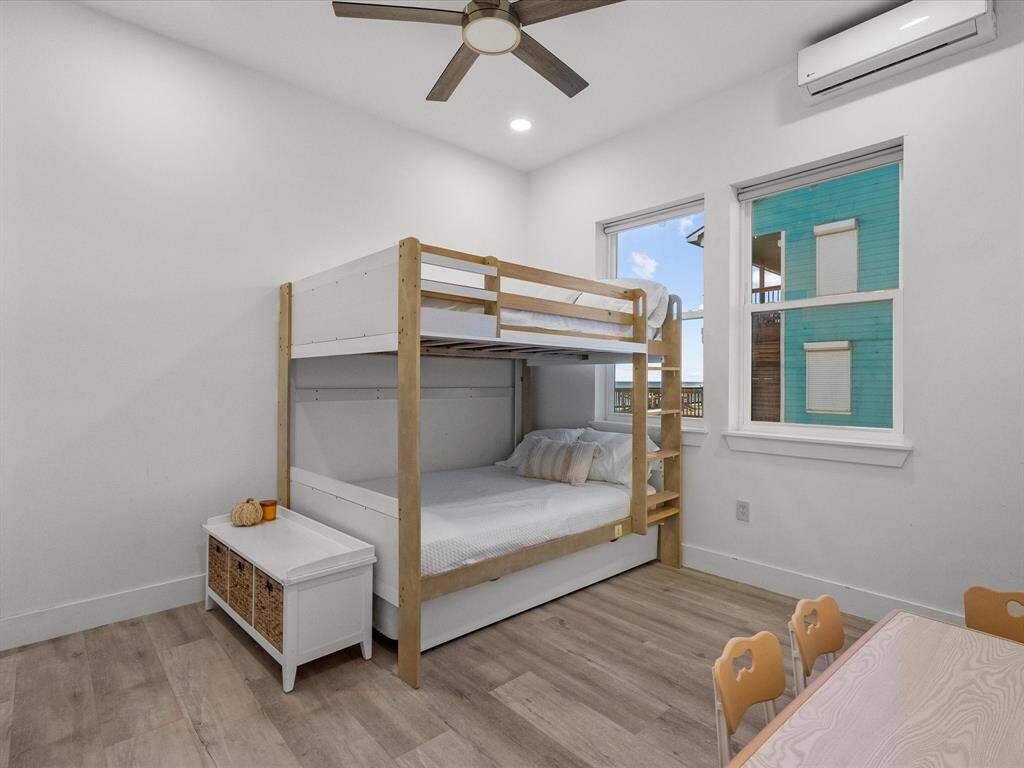 This bright bedroom features a modern bunk bed, a small play table with chairs, and a storage bench. It has hardwood floors, recessed lighting, and plenty of natural light from two large windows.