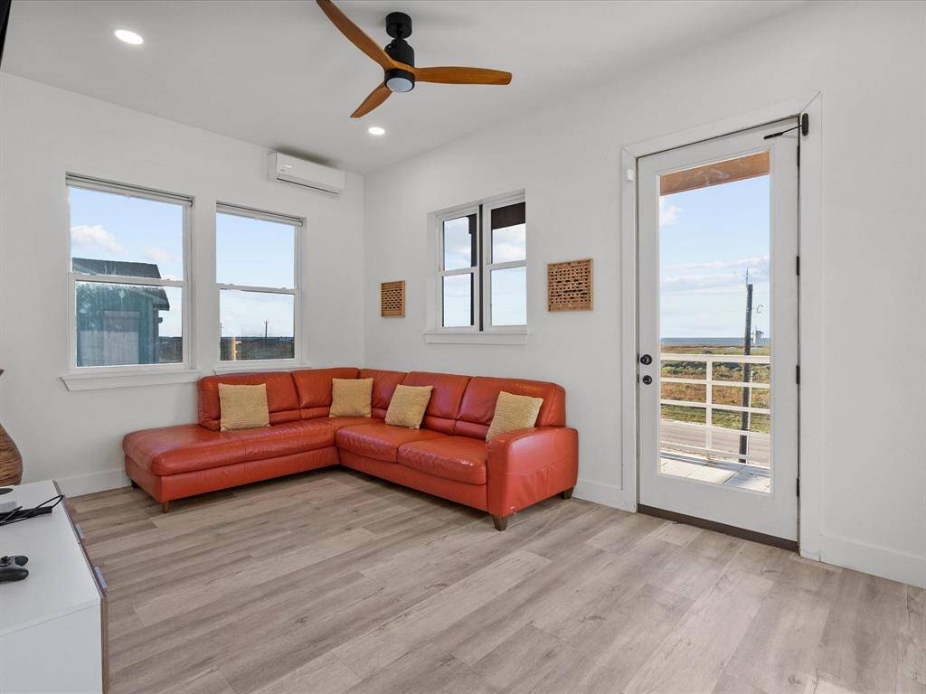 This cozy living room features a bright, spacious layout with a modern orange sectional sofa, large windows for natural light, wood flooring, and a ceiling fan. A door opens to an outdoor view, enhancing the airy feel.