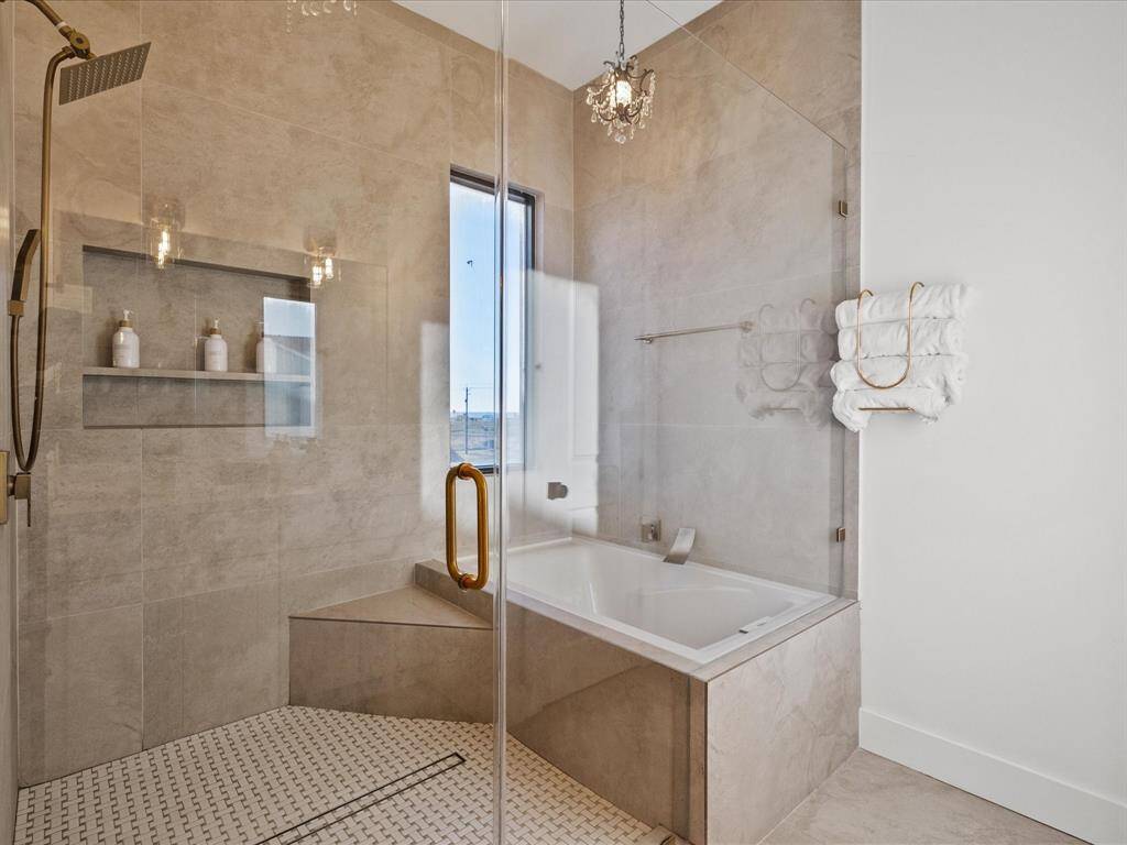This bathroom features a modern, spacious design with a glass-enclosed shower, a luxurious soaking tub, and elegant tile work. Natural light streams through a window, complementing the sophisticated chandelier and adding to the serene ambiance.