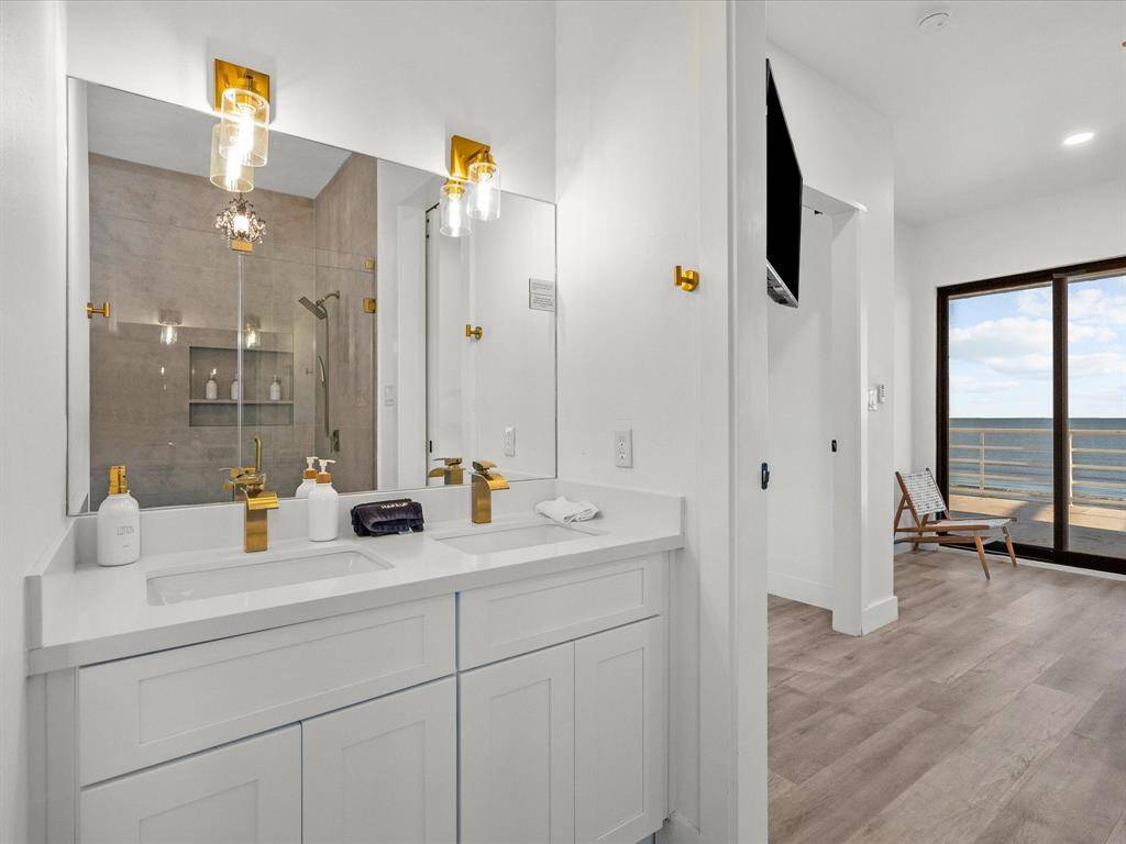 This bathroom features a modern double vanity with gold fixtures and glass pendant lighting. It opens to a bright room with hardwood floors and a large sliding glass door offering stunning ocean views.