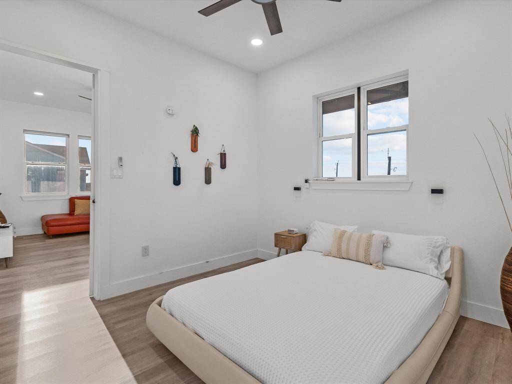 This photo shows a bright, minimalist bedroom with a modern design. It features a low platform bed, light wood flooring, and white walls. A window offers natural light, and the room connects to another space with more windows and a red sofa. Ceiling lights and decor add a cozy touch.