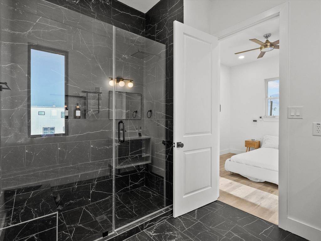This photo shows a modern bathroom with sleek black marble walls and flooring, featuring a glass-enclosed shower. It opens into a bright bedroom with light wood floors, a ceiling fan, and a large window for natural light.