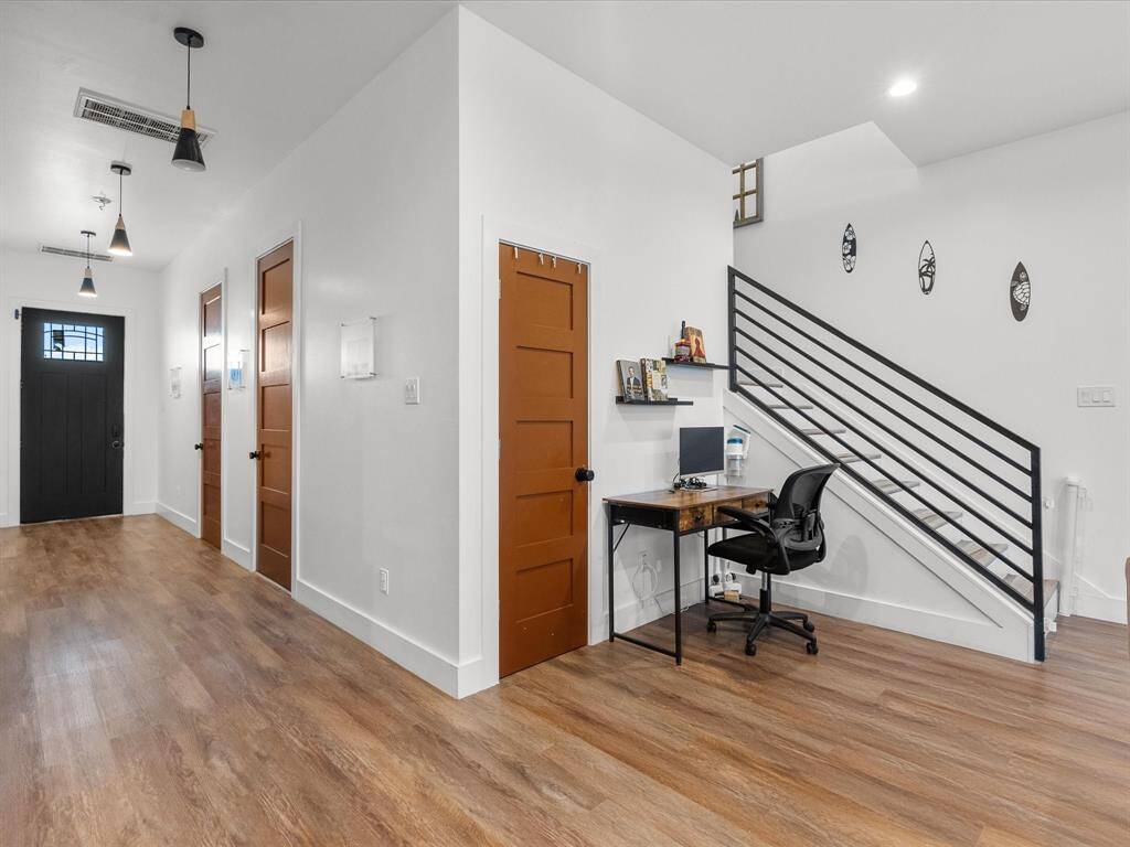 This photo showcases a modern entryway with wood flooring, white walls, and brown doors. It includes a small workspace under a staircase with a sleek black railing. The area is well-lit with pendant lights and has a clean, minimalist aesthetic.