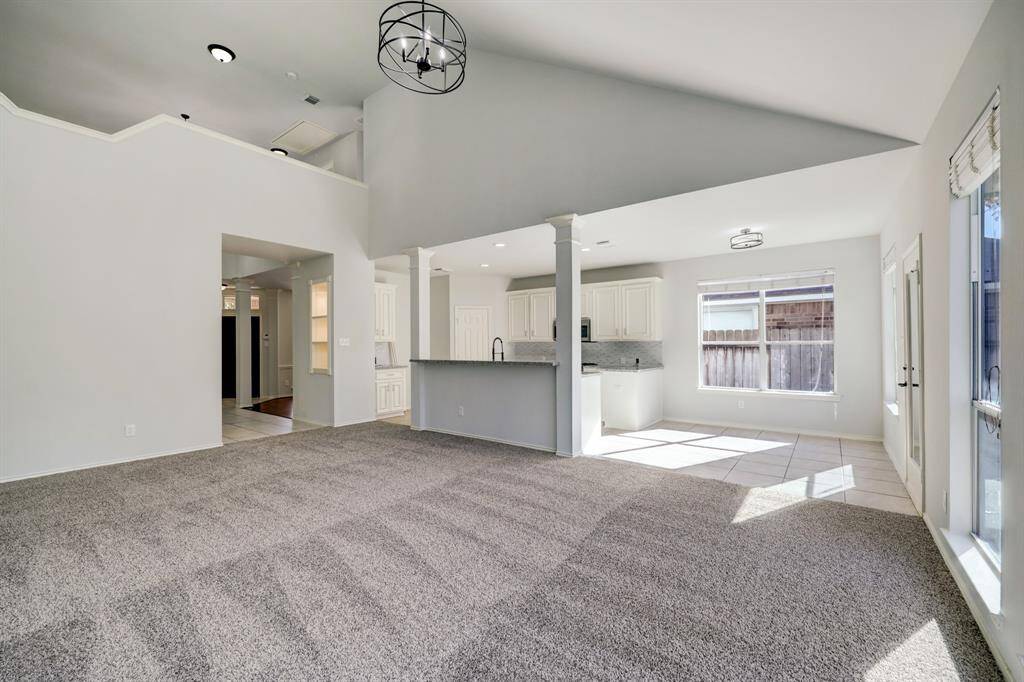 This photo shows a spacious, open-concept living area with high ceilings and ample natural light. It features a modern kitchen with white cabinetry and a breakfast bar, connecting seamlessly to a carpeted living space. Large windows and neutral tones create a bright, airy atmosphere.