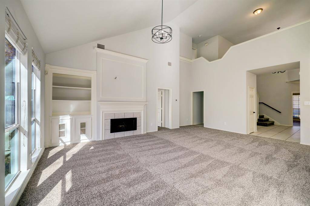 This spacious living room features a high ceiling and abundant natural light. It includes a modern chandelier, built-in shelving, and a fireplace. The neutral carpet complements the light walls, and there is easy access to other areas of the home through adjacent doorways.