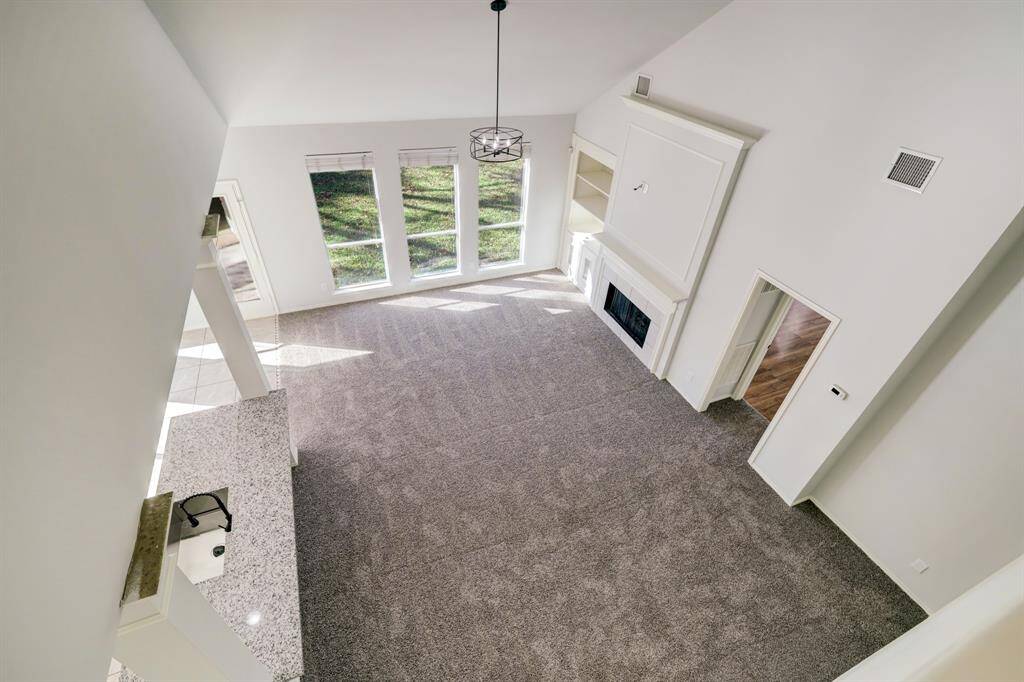 This spacious living room features high ceilings and large windows that allow for plenty of natural light. It includes a cozy fireplace with built-in shelves, and there's an open view into the kitchen with a granite countertop. The neutral carpet and walls create a bright, inviting atmosphere.