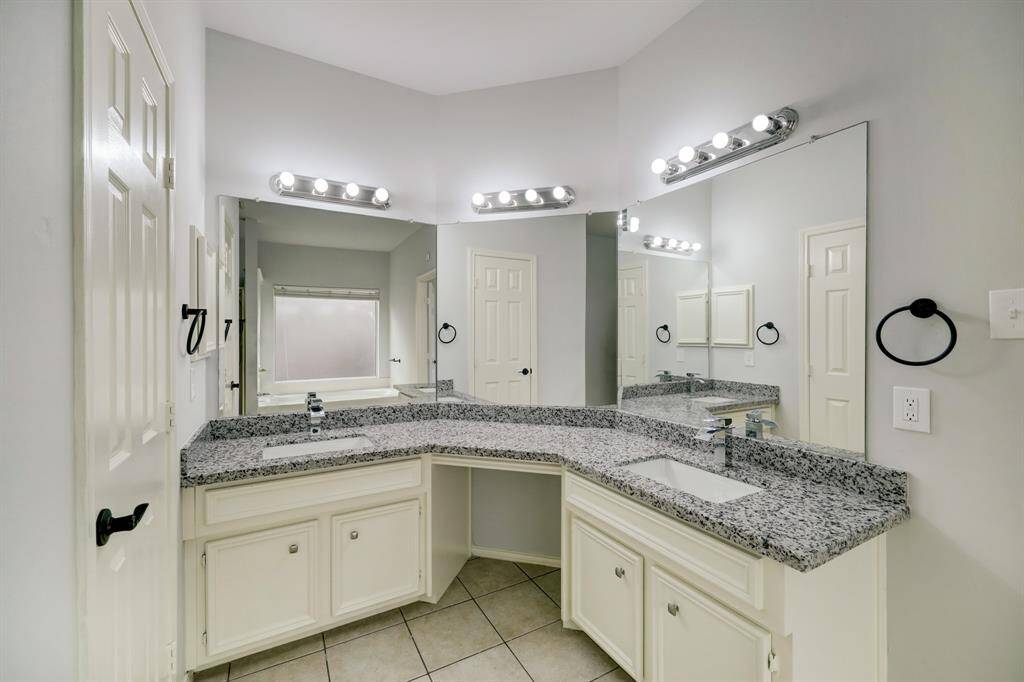 This bathroom features a spacious double vanity with granite countertops, modern fixtures, and large mirrors with ample lighting. The neutral tones and tiled floor add a clean, contemporary feel.