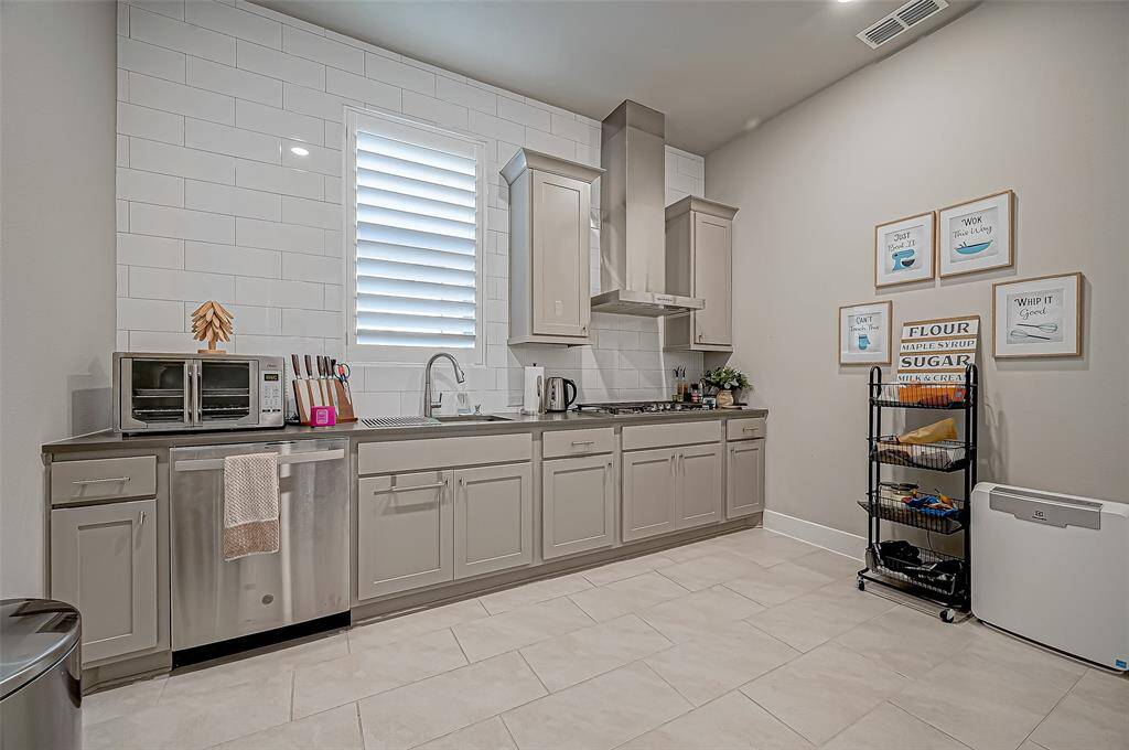 This modern hidden kitchen features sleek gray cabinetry, stainless steel appliances, and a stylish tile backsplash. It includes a built-in oven, dishwasher, and ample counter space, complemented by wall art and functional storage solutions.