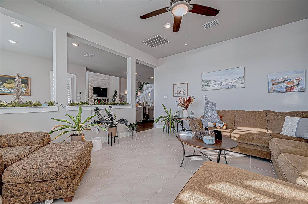 Bright and spacious living room with neutral tones, featuring a large sectional sofa, ceiling fan, and tile flooring. Open layout leads to a modern kitchen, enhanced by indoor plants and tasteful decor.