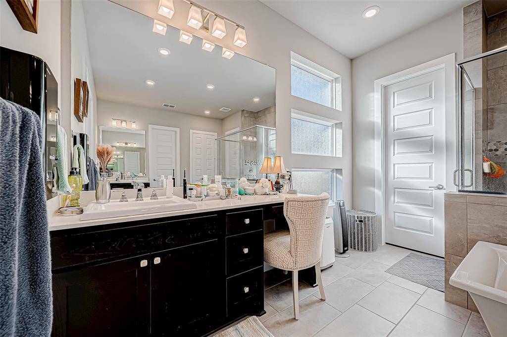 Bright and spacious bathroom featuring a dual-sink vanity, ample counter space, and modern lighting. The room includes a large mirror, a stylish chair, and a glass-enclosed shower. Natural light pours in through the high windows, enhancing the clean, contemporary design.