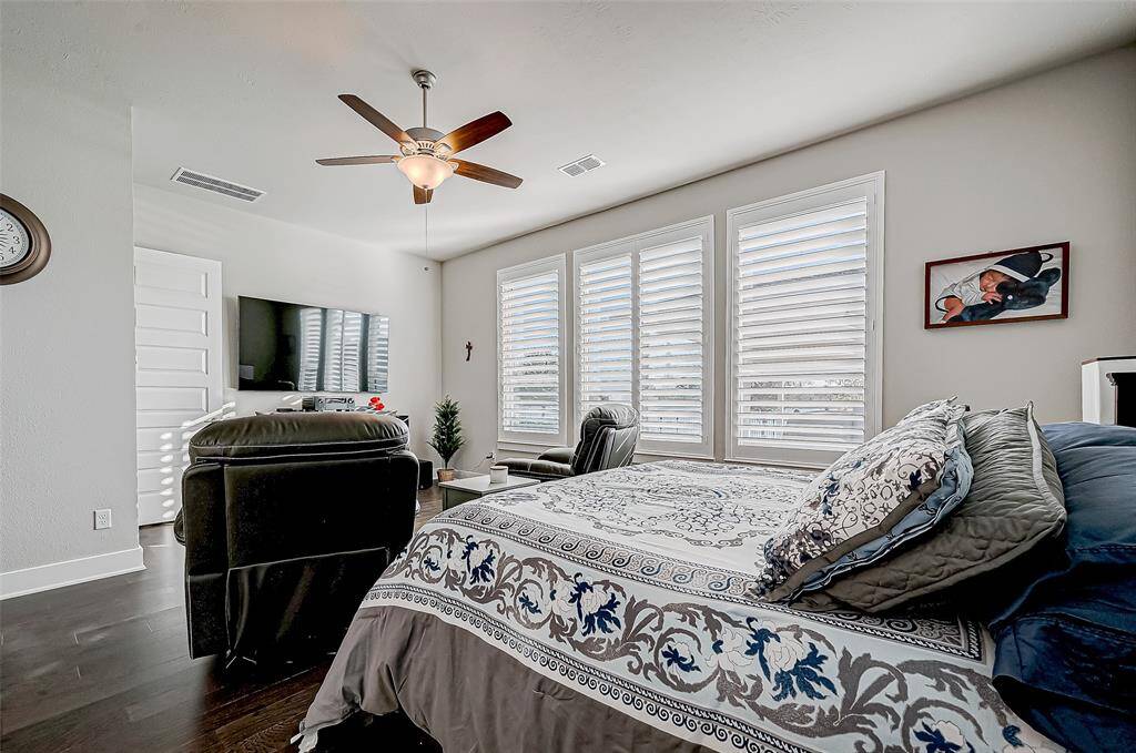 This spacious bedroom features large windows with shutters, a ceiling fan, and hardwood flooring. It includes a cozy sitting area with a TV, offering plenty of natural light and a comfortable, modern vibe.