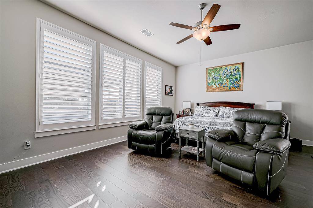 This cozy bedroom features large windows with shutters, allowing natural light to fill the space. It includes a comfortable bed, two plush chairs, and a ceiling fan. The hardwood floors and neutral walls add a touch of elegance.