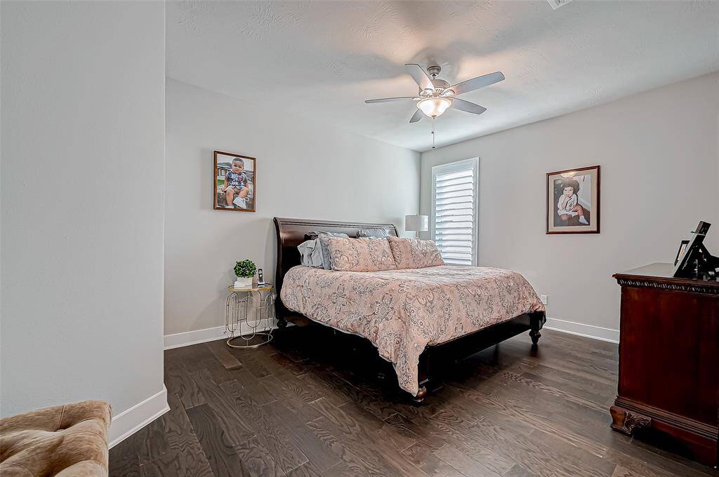 This bedroom features a cozy atmosphere with a large bed, dark wood flooring, and a ceiling fan. It has a side table with a lamp, decorative wall art, and a window with shutters, offering a blend of comfort and style.