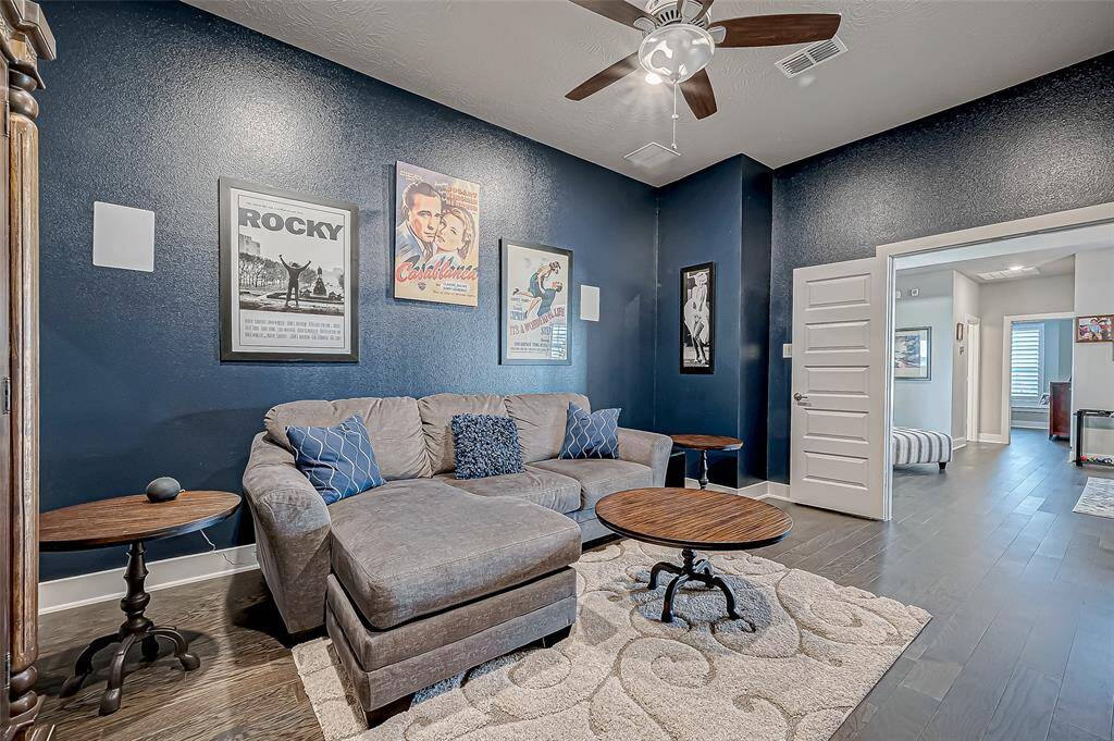 This cozy living space features dark blue walls adorned with classic movie posters, a comfortable sectional sofa, and stylish round tables. The ceiling fan adds a modern touch, and the adjacent room offers additional living space or a bedroom. Perfect for relaxation and entertainment.