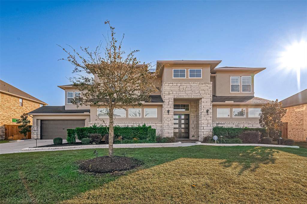 This is a modern two-story home with a clean, contemporary facade featuring stone and stucco elements. It has a well-maintained lawn and a four-car garage. Large windows provide plenty of natural light, and the entrance is inviting with landscaped greenery.