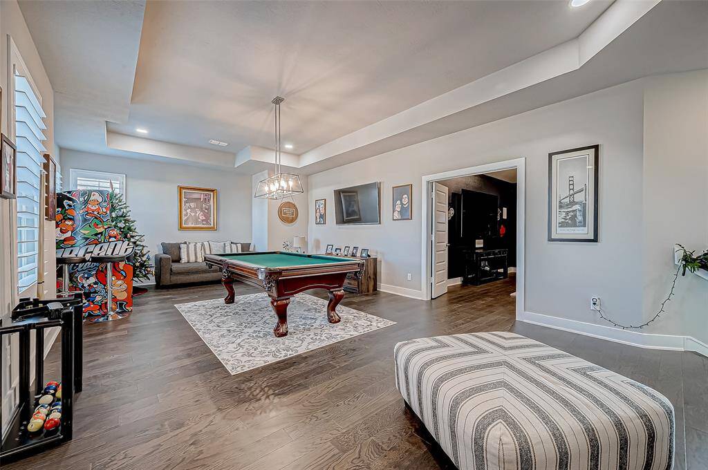 This photo shows a stylish game room featuring a pool table on a decorative rug. It has hardwood floors, a cozy seating area, and vibrant artwork. The room is well-lit with recessed and pendant lighting, offering a modern and inviting atmosphere.