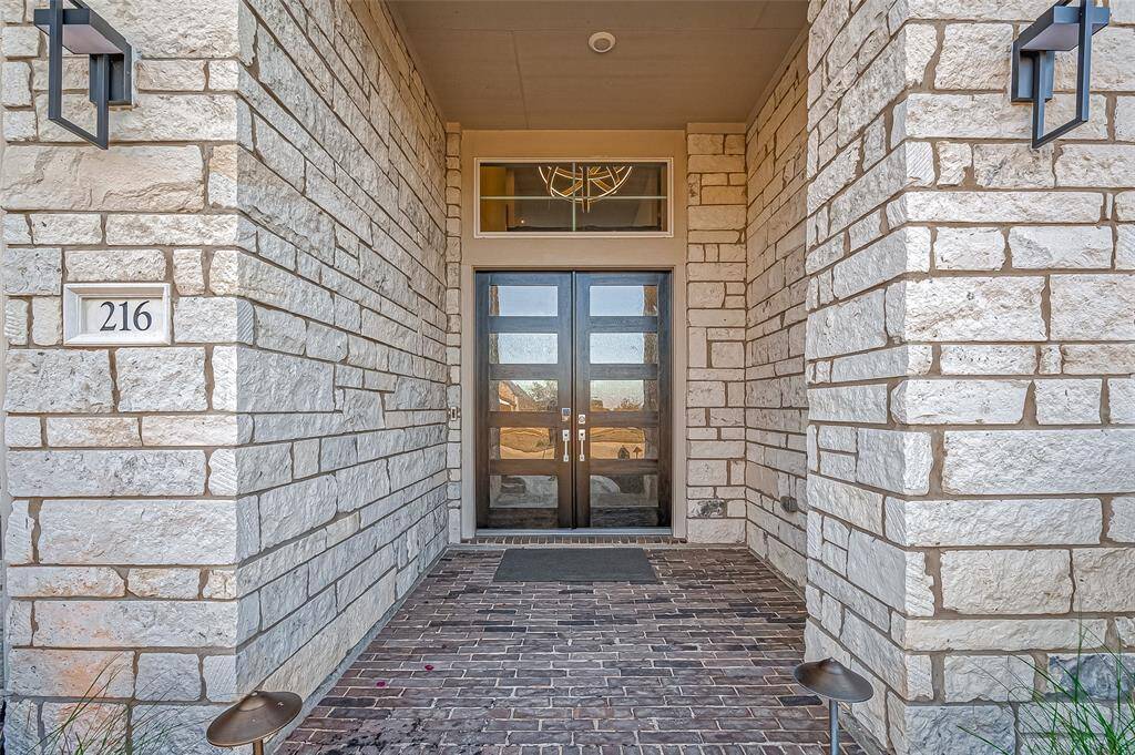 This photo showcases a modern front entrance with a double door set against a stylish stone facade. The entryway is framed by elegant lighting fixtures and features a textured brick pathway.
