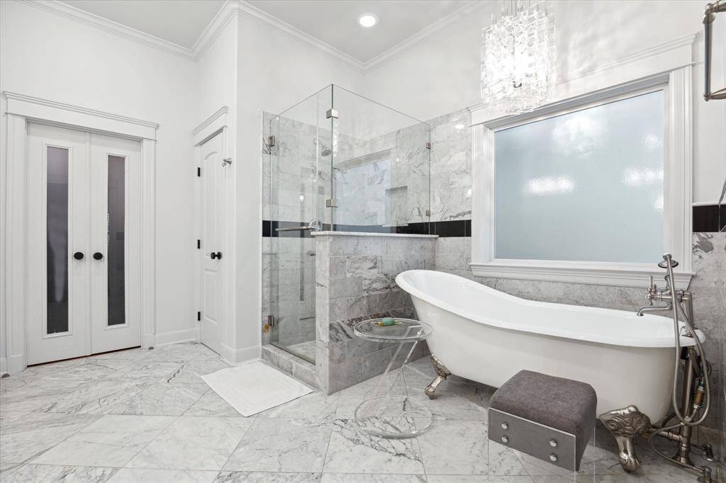 The primary bathroom features elegant marble flooring and walls, a spacious glass-enclosed shower, and a luxurious clawfoot tub. It’s bright and modern, with a chic chandelier and frosted window for privacy.