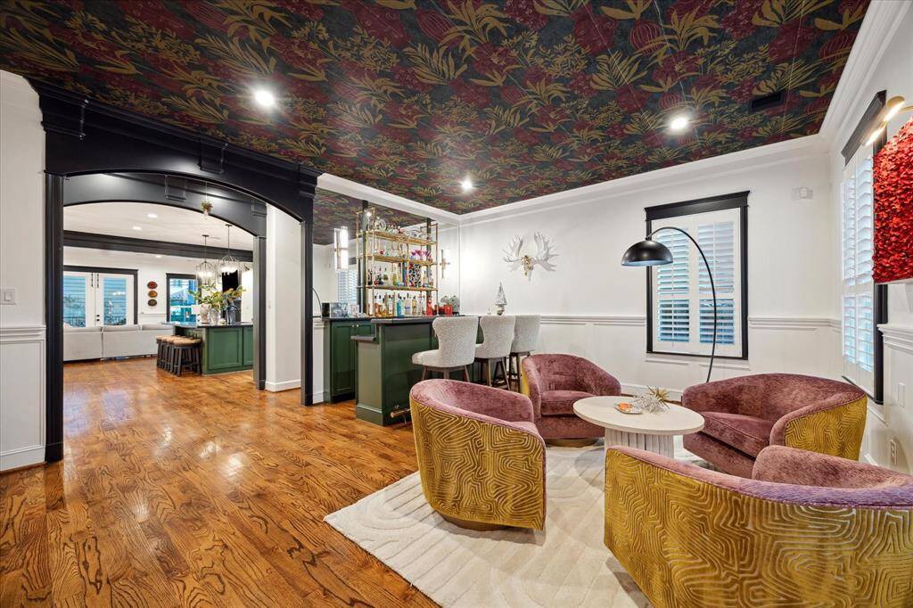 This vibrant living area features a stylish bar with green cabinetry, rich hardwood floors, and bold patterned ceilings. Cozy seating with plush pink and gold chairs creates a welcoming atmosphere, perfect for entertaining. The open design seamlessly connects to rest of the first floor living spaces