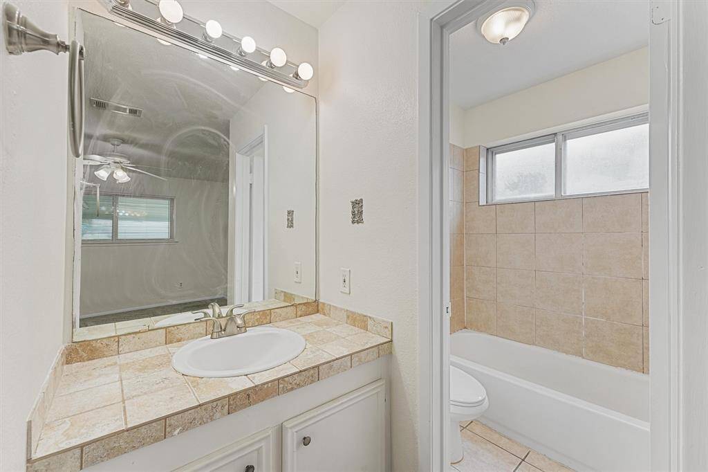 This bathroom features a tiled vanity with a large mirror and bright lighting, leading to a bathtub with a tiled surround and frosted windows for privacy.