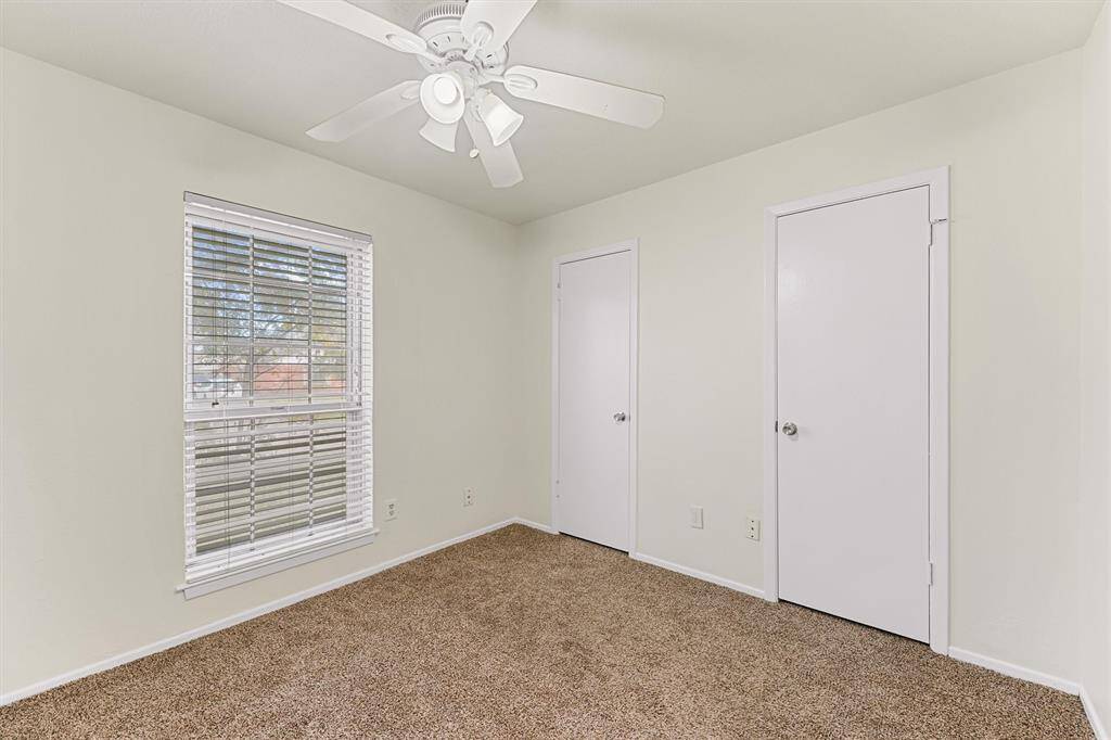 This is a cozy, carpeted bedroom with neutral walls, featuring a ceiling fan for comfort. It has a window with blinds for natural light and two closets.