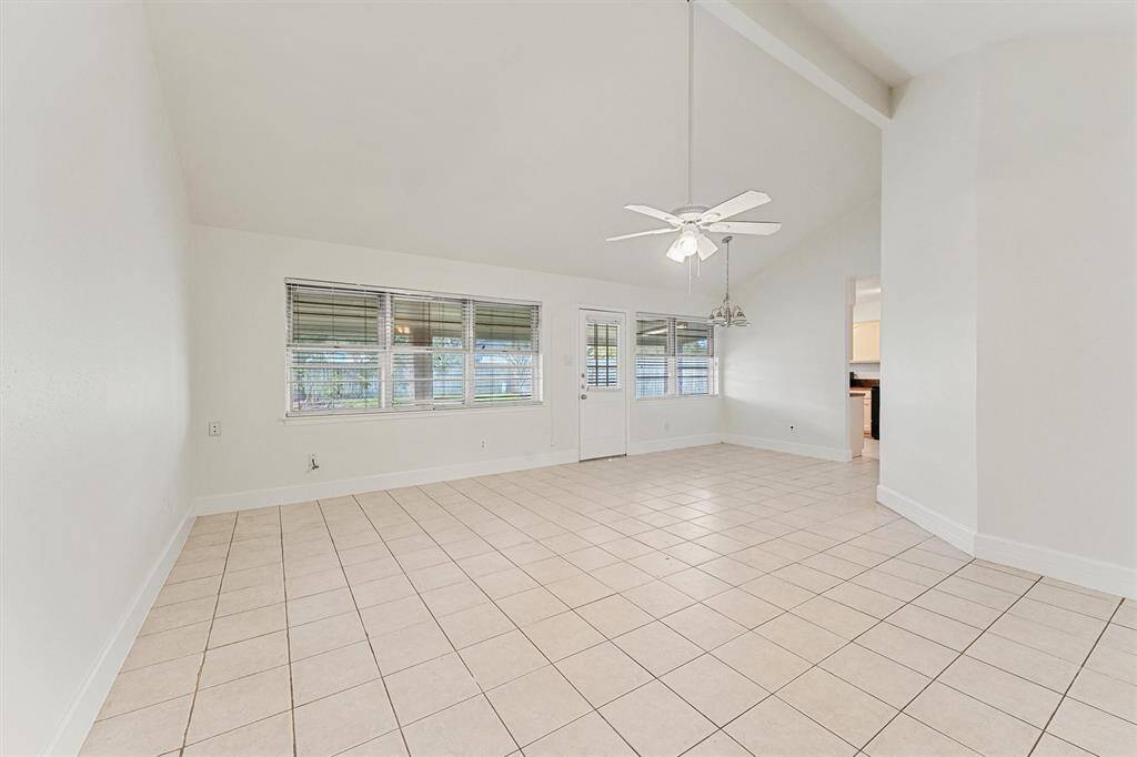This spacious room features a high vaulted ceiling, tile flooring, and ample natural light from large windows. A ceiling fan and light fixtures add functionality, and the neutral tones offer a versatile space for any decor style.