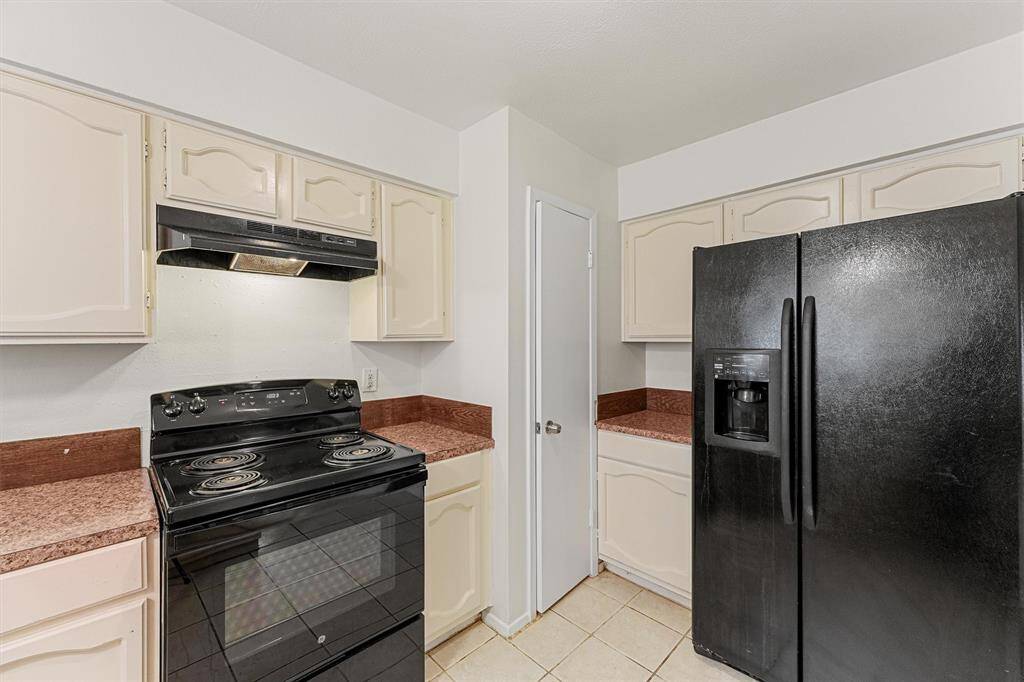 The kitchen features light cabinetry and countertops, a black electric stove, and a matching black refrigerator. The space is compact and functional, with tiled flooring and a simple design.