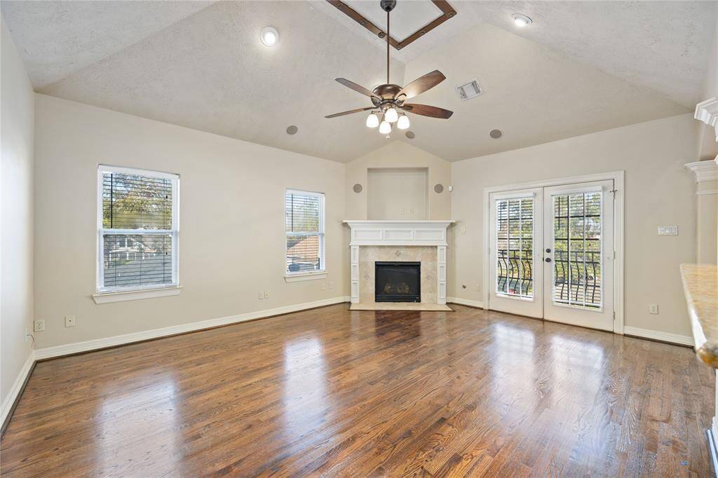 This spacious living area features hardwood floors, a cozy fireplace, and high ceilings with recessed lighting. It opens to a modern kitchen with stainless steel appliances and elegant wooden cabinetry. The room is brightened by natural light from large French doors leading to an outdoor space.