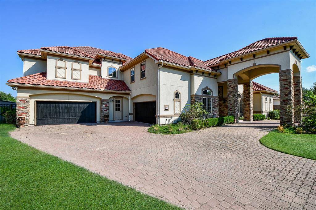 This is a spacious two-story home featuring a stucco finish with stone accents, a terracotta tile roof, and an arched entryway. It includes a large driveway and an attached four-car garage. The property has well-maintained landscaping and offers a luxurious curb appeal.