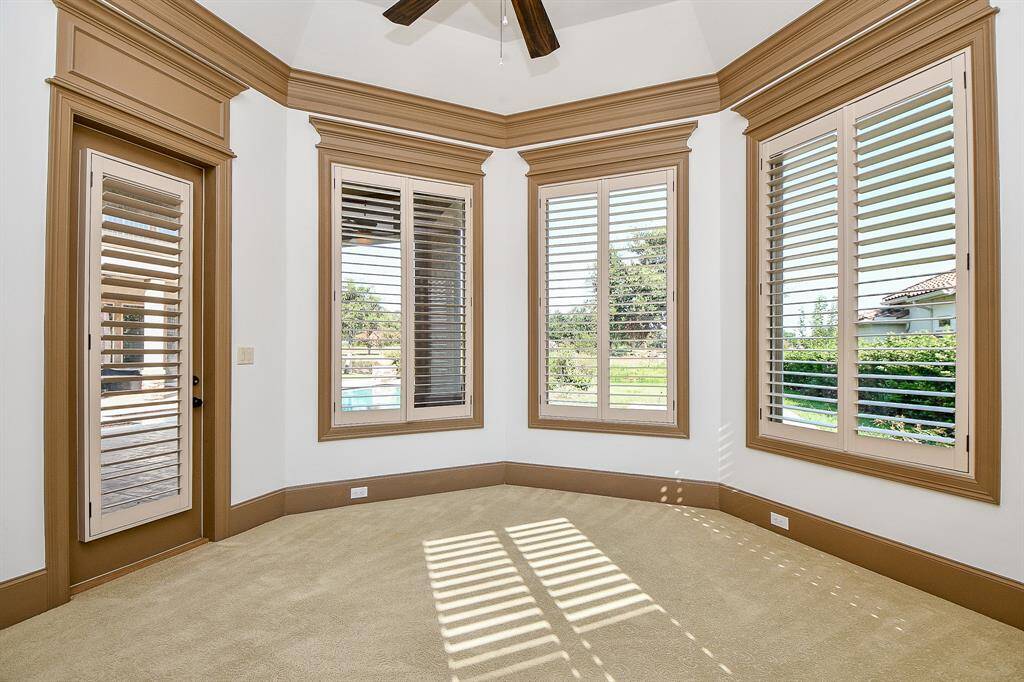 This is part of the primary suite featuring elegant crown molding, a high ceiling with exposed beams, plantation shutters on multiple windows providing ample natural light, and plush carpeting. It offers a view of the outdoors and has direct access to another area through a door with louvered shutters.