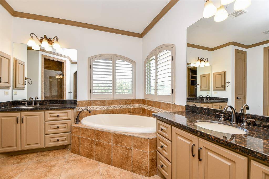 This is a spacious primary bathroom featuring a large corner bathtub with tile surround, dual vanity with granite countertops, ample cabinetry, tiled flooring, and two windows providing natural light.