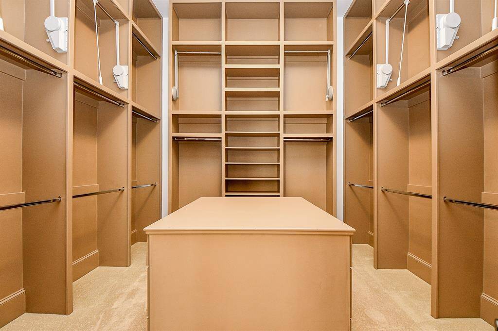 This is a spacious primary bedroom walk-in closet with ample built-in shelves, drawers, and hanging rods for organized storage. A central island provides additional countertop space for accessories or folding clothes. The closet is well-lit with several light fixtures.