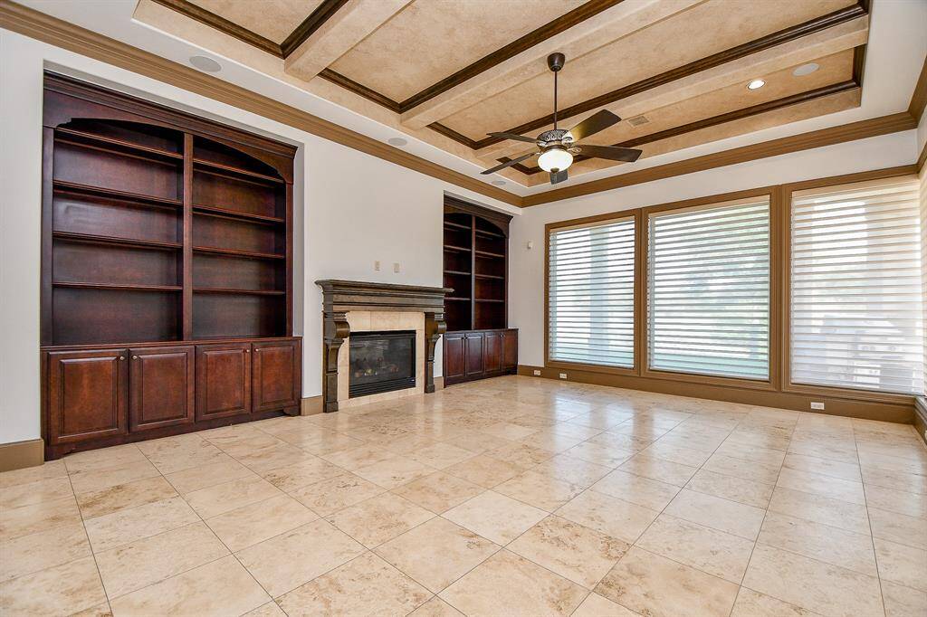 This is a spacious living room with tiled flooring, featuring a large built-in wooden bookshelf, an elegant fireplace, and natural light from windows with blinds. The room has a coffered ceiling with a central fan, adding a touch of luxury.