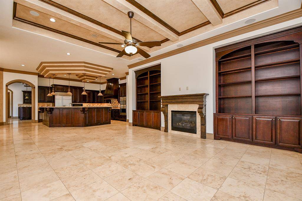 This is a spacious, open-concept living space with a large kitchen featuring wood cabinets and a central island. There's a cozy fireplace and built-in shelving, perfect for storage and display. The room boasts high ceilings with recessed lighting and a ceiling fan, and is finished with elegant tile flooring.