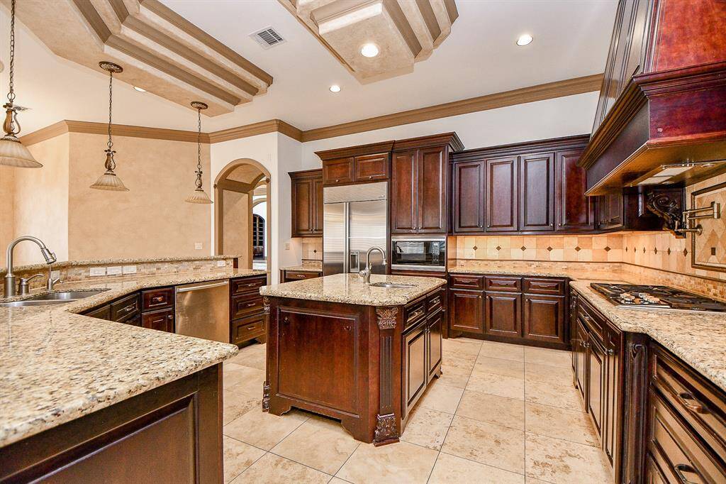 Spacious kitchen with rich, dark cabinetry, granite countertops, a central island, stainless steel appliances, and elegant hanging light fixtures. Features include a decorative ceiling with recessed lighting and a tiled floor.