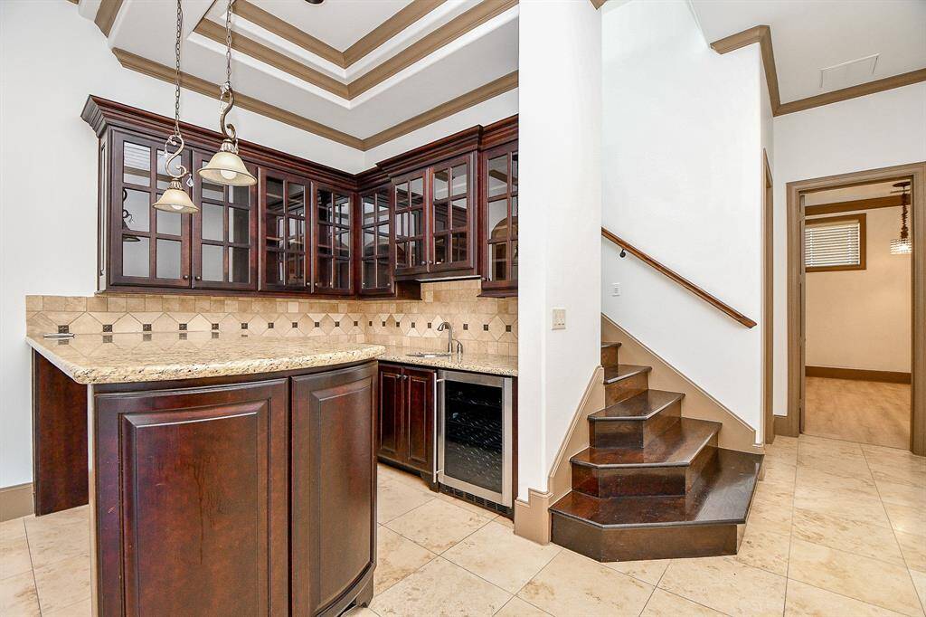 This photo features a kitchen with dark wood cabinetry, granite countertops, and a wine fridge, complemented by a tray ceiling with molding details. To the right is a staircase leading to another level and a doorway to an adjacent room. The space is finished with tile flooring.