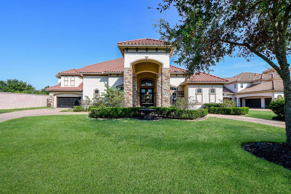 This is a spacious two-story home featuring a Mediterranean style with a stucco exterior, a tile roof, and an arched entryway. It has a well-manicured lawn, an attached garage, and is situated in a suburban neighborhood with similar upscale homes.