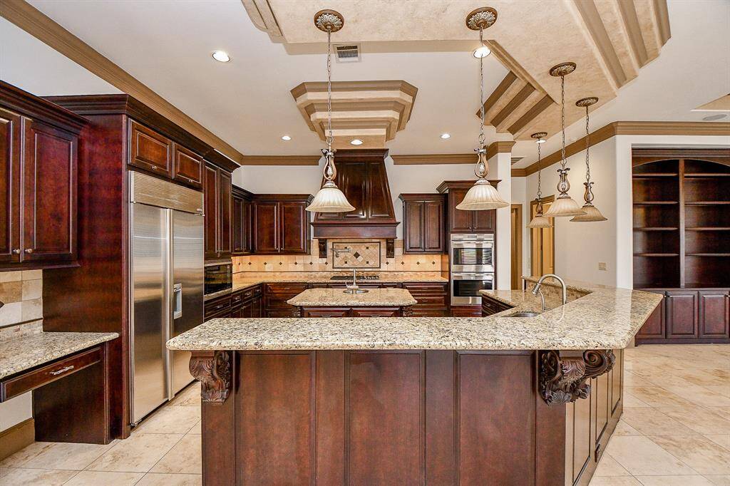 This is a spacious kitchen with rich, dark wood cabinetry, granite countertops, and high-end stainless steel appliances. It features a large center island, pendant lighting, and an adjacent built-in bookshelf area, all under an elegant tray ceiling.