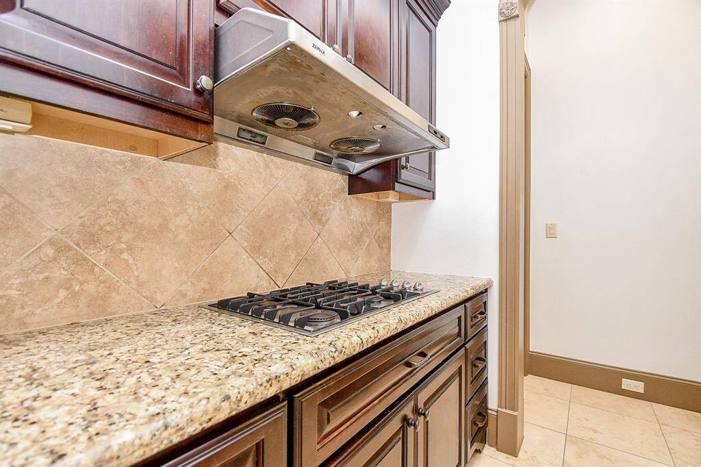 This kitchen features rich wooden cabinetry, a sleek gas cooktop, and granite countertops, complemented by a tiled backsplash and a stainless-steel vent hood.