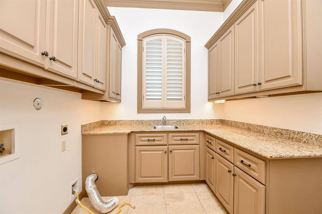 This is a utility room featuring cream-colored cabinetry with traditional styling, complemented by granite countertops. A small, arched window with shutters provides natural light. The room includes a built-in sink. There's an exposed flexible duct for Dryer hookup.