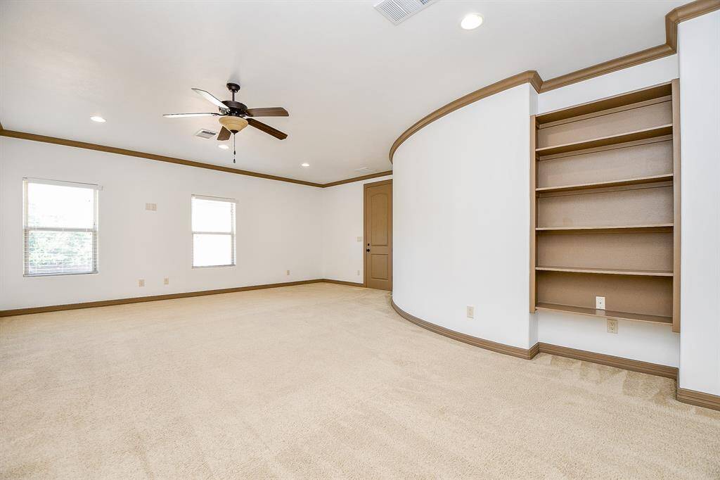 This is a spacious room with neutral carpeting, two windows allowing for natural light, a ceiling fan for comfort, and built-in shelving for storage or display.