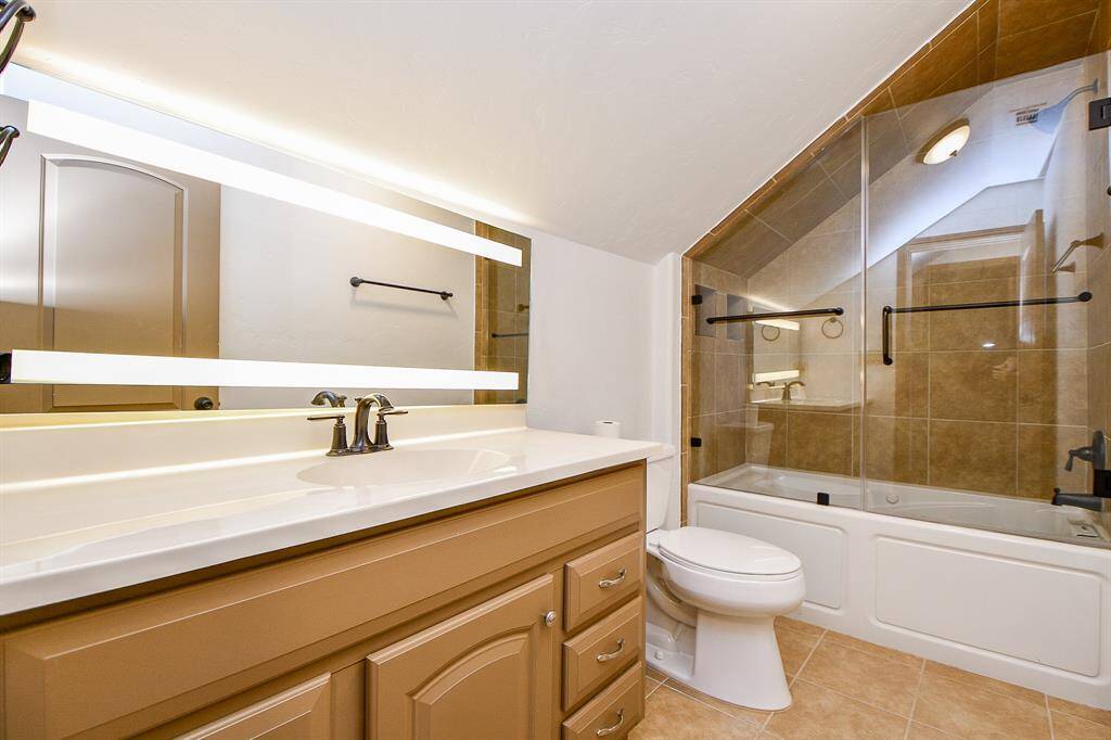 This is a well-lit bathroom featuring a large mirror, a vanity with ample storage, dual sink faucets, and a full bathtub with a glass enclosure. The room has a neutral color palette with beige wall tiles.