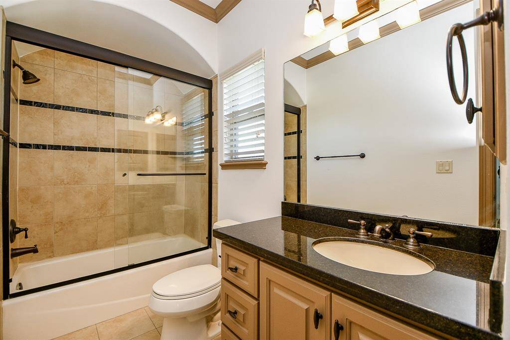 This is a modern bathroom featuring a tiled shower-tub combo with glass doors, a single vanity with ample storage and granite countertop, and a toilet. The space is well-lit, with a window providing natural light.
