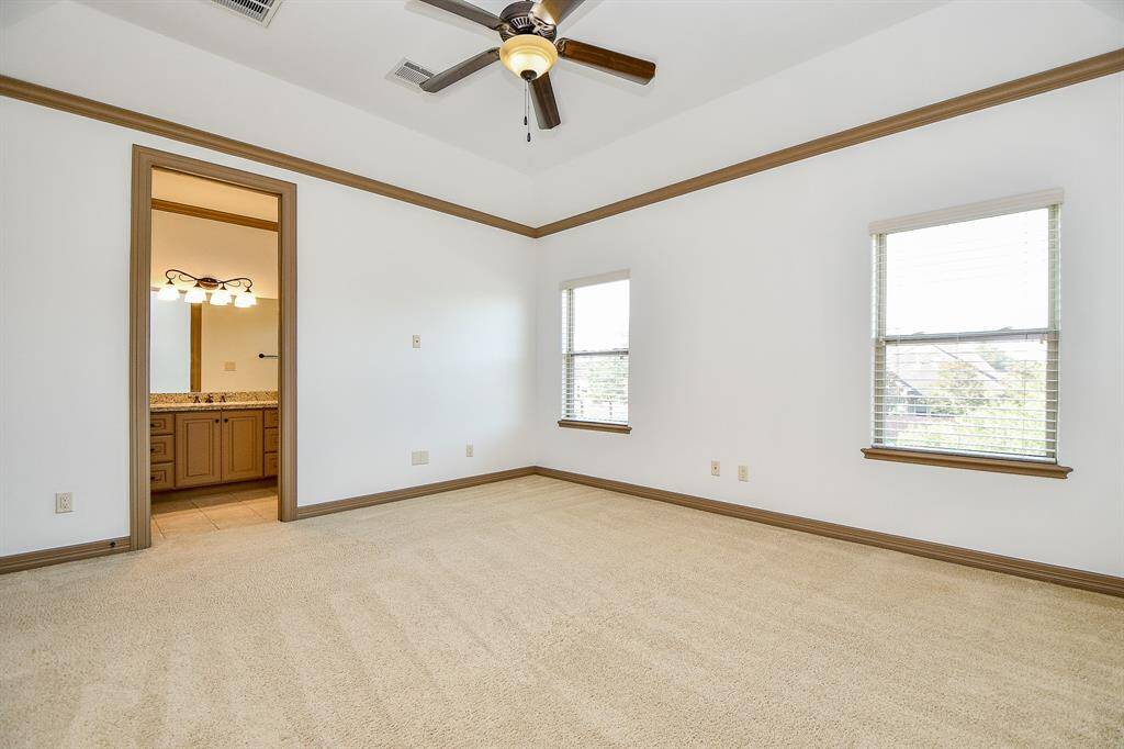 This is a spacious, well-lit bedroom with beige carpeting, two large windows with blinds, a ceiling fan, and an en suite bathroom visible through an open doorway.