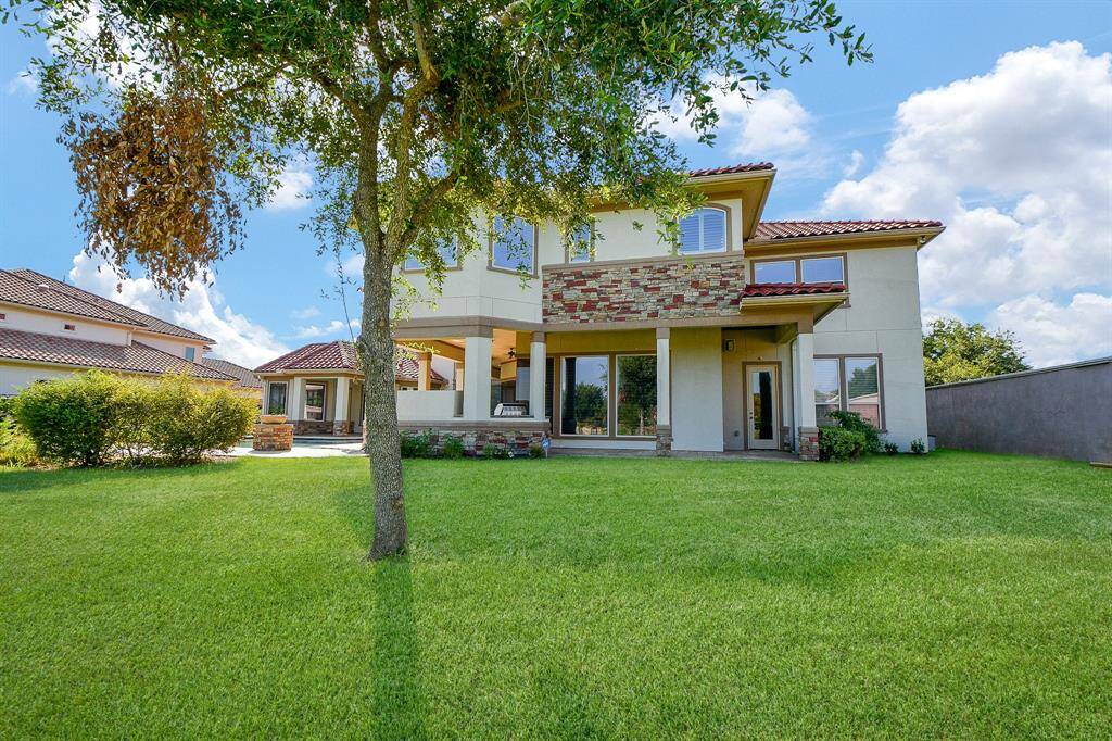 This is a modern two-story home with a stucco and brick facade, featuring a balcony, large windows, and a spacious lawn. It is situated in a sunny, suburban setting with clear skies above.