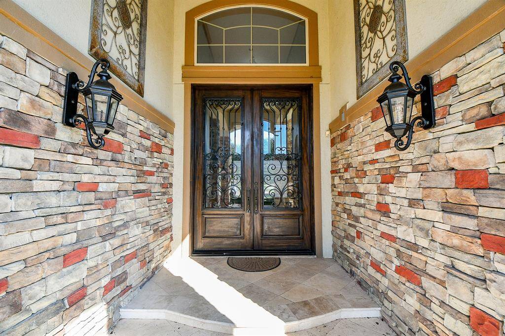 This photo showcases an elegant home entrance featuring a beautifully crafted wrought-iron double door flanked by classic wall lanterns, all set against a charming stone veneer facade.