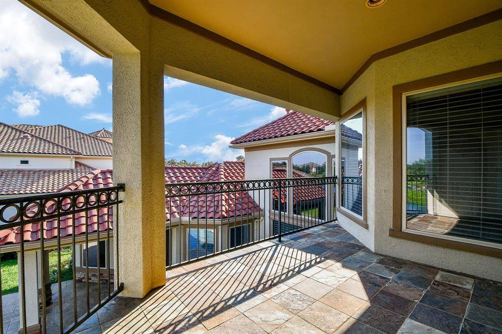 This is a spacious covered balcony with a picturesque view, featuring elegant tile flooring and a classic wrought-iron railing that offers both safety and style. The balcony overlooks a serene neighborhood with similar upscale homes, and the area is bathed in natural light.